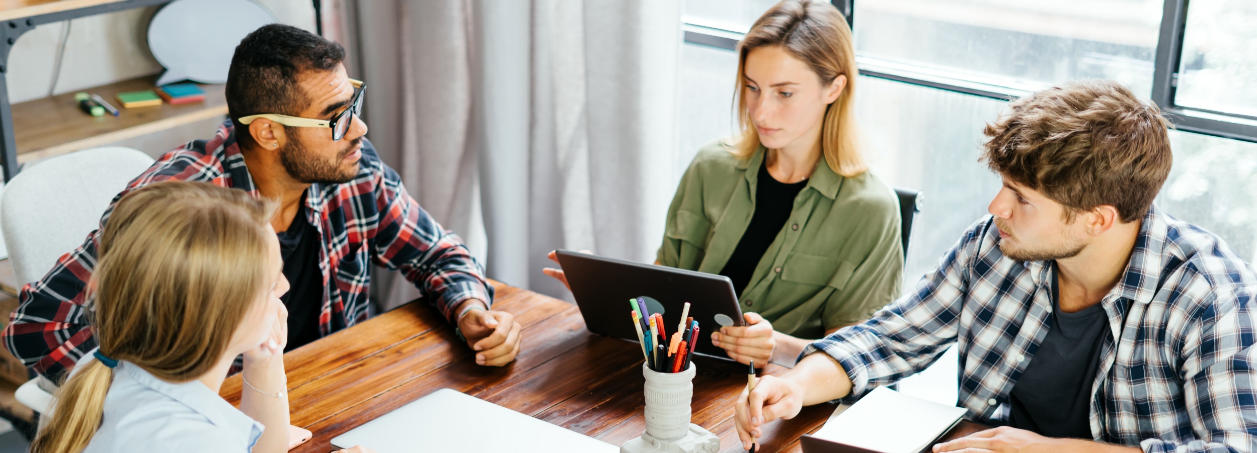 Vier Personen sitzen an einem Holztisch und scheinen in einer Besprechung oder Zusammenarbeit vertieft zu sein.

Auf dem Bild sind vier Personen zu sehen, um einen großen, dunkelbraunen Holztisch versammelt. Links sitzt eine junge Frau mit blonden Haaren, die nachdenklich in die Ferne blickt. Neben ihr sitzt ein Mann mit dunkler Haut, Bart und Brille, der ebenfalls konzentriert wirkt und etwas erklärt. Gegenüber sitzen eine Frau, die ein Tablet in den Händen hält und dieses zu präsentieren scheint, und ein Mann mit lockigem Haar, der Notizen macht. Auf dem Tisch befinden sich ein Laptop, ein Stifthalter in Kameraoptik mit bunten Stiften, ein Notizbuch und andere Arbeitsutensilien. Im Hintergrund ist ein Fenster mit Vorhängen und ein Regal zu sehen. Die Atmosphäre wirkt professionell, konzentriert und kollaborativ. Das Bild vermittelt den Eindruck eines kreativen Teams, das gemeinsam an einem Projekt arbeitet.
