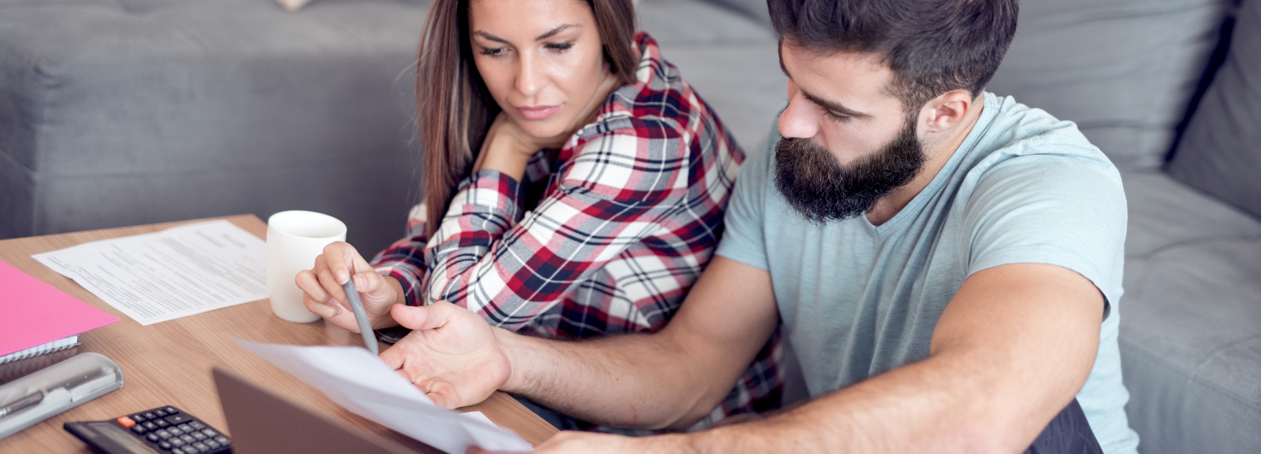 Ein junges Paar sitzt an einem Tisch und scheint konzentriert über Dokumente und Finanzen zu sprechen.

Das Bild zeigt ein junges Paar, das an einem hellbraunen Holztisch sitzt. Die Frau trägt ein rot-weiß kariertes Hemd und hat lange, braune Haare mit blonden Strähnen. Sie stützt ihren Kopf mit der Hand ab und scheint konzentriert auf die Dokumente vor ihr zu schauen. Der Mann neben ihr hat einen Bart und trägt ein hellblaues T-Shirt. Er hält ein Blatt Papier in der Hand und deutet mit der anderen Hand darauf, als würde er etwas erklären. 

Auf dem Tisch befinden sich mehrere Dokumente, ein pinkfarbener Notizblock, ein Taschenrechner, ein Bleistift und eine weiße Kaffeetasse. Im Hintergrund ist ein graues Sofa zu sehen. Die Atmosphäre wirkt ernst und konzentriert, möglicherweise diskutieren sie über ihre Finanzen oder andere wichtige Angelegenheiten. Das Licht ist hell und natürlich, was dem Bild eine ruhige und häusliche Note verleiht.