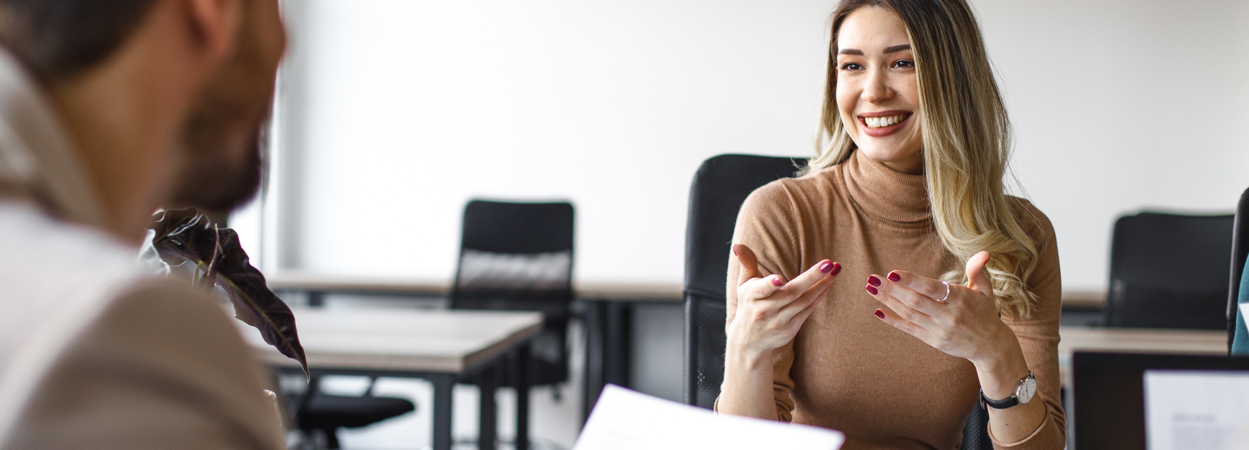 Das Bild zeigt ein junges, lächelndes Frau, das in einem professionellen Umfeld gestikulierend spricht, während sie von einer schlecht definierten Person gegenüber zu sitzen scheint.

Das Bild zeigt eine Frau, die in einem Büro oder Konferenzraum sitzt. Sie hat langes, blondes Haar, das über ihre Schultern fällt, und trägt einen braunen Rollkragenpullover. Ihr Gesicht ist leicht zur Seite gedreht und sie lächelt herzlich, wobei ihre Zähne sichtbar sind. Ihre Augen sind wach und scheinen auf eine Person zuzublicken, die außerhalb des Bildausschnitts liegt. Ihre Hände sind erhoben und sie gestikuliert mit ihnen, als ob sie etwas erklärt oder mit Begeisterung erzählt. Ihre Fingernägel sind in einer dunklen Farbe lackiert. Sie trägt einen Ring an ihrer linken Hand und eine Armbanduhr am linken Handgelenk.

Im Vordergrund ist unscharf eine Pflanze mit dunklen Blättern zu sehen, die sich von links ins Bild schiebt. Hinter der Frau sieht man unscharf zwei schwarze Bürostühle und einen Tisch mit einer glatten, hellen Oberfläche. Weiter hinten, im Hintergrund, sind eine weiße Wand und weitere unscharfe Büromöbel angedeutet, was auf eine professionelle Arbeitsumgebung hindeutet. Die Beleuchtung im Raum ist hell und gleichmäßig, was eine klare und offene Atmosphäre schafft.

Die Stimmung des Bildes ist positiv und energiegeladen. Das Lächeln und die Gestik der Frau deuten auf ein engagedes und angenehmes Gespräch hin, möglicherweise eine Präsentation, eine Besprechung oder ein Vorstellungsgespräch, bei dem sie sich wohlfühlt und ihre Ideen lebhaft vermittelt. Die Unschärfe im Vordergrund und Hintergrund lenkt den Fokus auf die Frau und ihre Interaktion, während die gesamte Szene Professionalität und dynamische Kommunikation ausstrahlt.