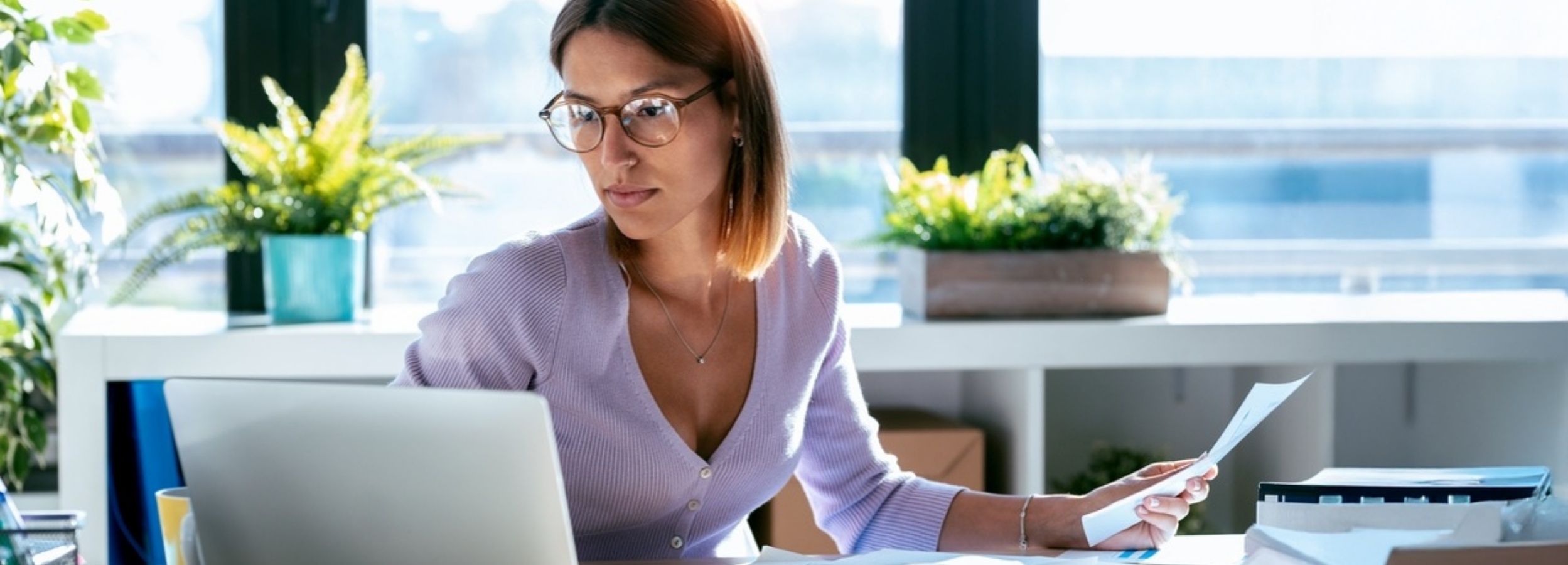 Das Bild zeigt eine nachdenkliche Frau, die an ihrem Schreibtisch arbeitet, umgeben von Pflanzen und hellem Tageslicht. 

Auf dem Bild ist eine junge Frau mit schulterlangem, dunkelbraunem Haar und Brille zu sehen, die an einem Schreibtisch sitzt und sich auf ihren Laptop und einige Papiere konzentriert. Sie trägt einen hellvioletten Cardigan mit Knöpfen. Ihr Blick ist gesenkt, was auf Konzentration oder Nachdenklichkeit hindeutet. Sie hält ein Blatt Papier in ihrer rechten Hand in der Nähe ihres Gesichts, während ihre linke Hand auf dem Laptop ruht. Am Schreibtisch sind ein offener Laptop, verschiedene Dokumente, ein Stiftehalter mit Stiften und ein gelber Becher zu erkennen. Im Hintergrund ist ein Fenster mit hellem Tageslicht zu sehen, das den Raum beleuchtet. Auf dem Fensterbrett und einem Regal stehen mehrere grüne Pflanzen, die dem Arbeitsplatz eine lebendige und erfrischende Note verleihen. Die übergeordnete Atmosphäre ist ruhig und konzentriert, mit einer leichten Wärme, die durch das Sonnenlicht und die Pflanzen erzeugt wird. Die Stimmung scheint ruhig und geschäftig zu sein, was auf eine Frau hinweist, die in ihre Arbeit vertieft ist und eine angenehme Arbeitsumgebung genießt.