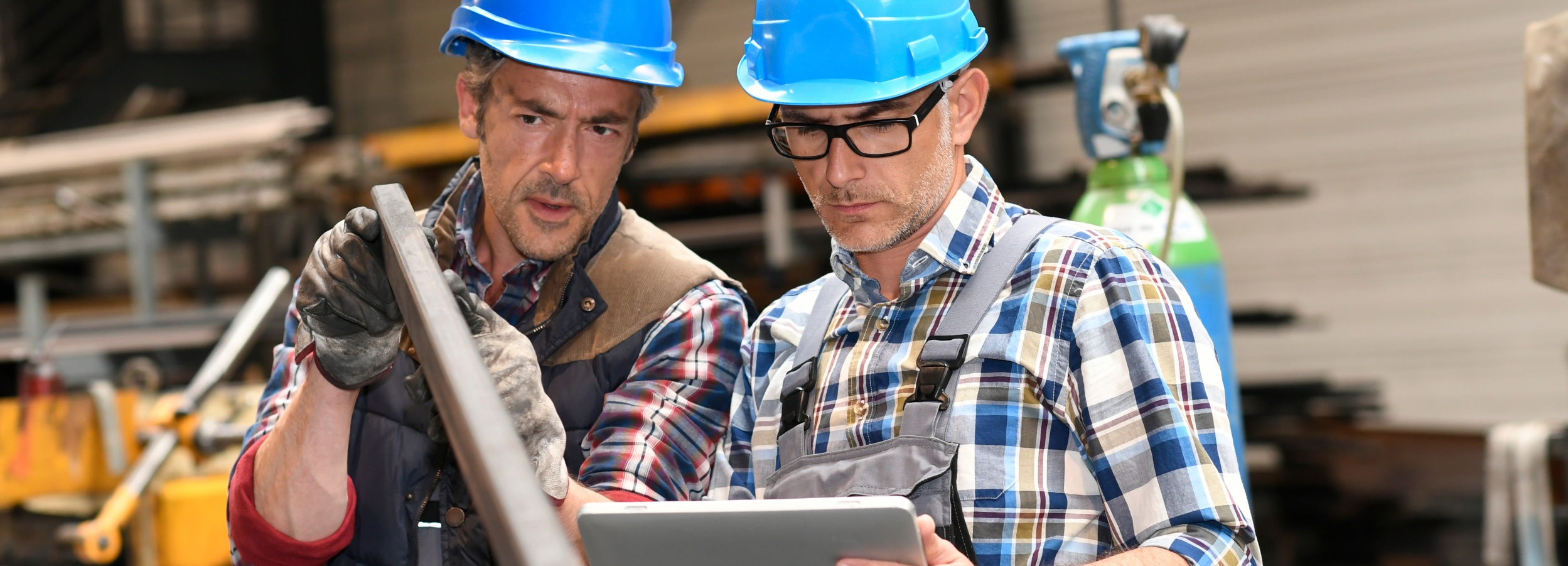Zwei Bauarbeiter mit blauen Schutzhelmen diskutieren und prüfen ein Metallteil, während sie ein Tablet benutzen.

Das Bild zeigt zwei Männer in einer industriellen Umgebung, wahrscheinlich einer Werkstatt oder Baustelle. Beide tragen blaue Schutzhelme und Arbeitskleidung. Der Mann links, der etwas älter aussieht und eine westenartige Arbeitsjacke über einem karierten Hemd trägt, hält mit beiden Händen ein langes, rechteckiges Metallprofil. Seine Hände stecken in schmutzigen Arbeitshandschuhen. Er hat schmutzverschmierte Wangen und einen ernsten, konzentrierten Gesichtsausdruck, während er auf das Metallstück blickt. Seine Augen sind leicht zusammengekniffen, so als ob er etwas genau beurteilt oder erklärt.

Der Mann rechts, der jünger wirkt und eine Latzhose über einem karierten Hemd trägt, blickt auf ein silbernes Tablet, das er in den Händen hält. Er trägt eine Brille mit schwarzem Rahmen und hat ebenfalls einen konzentrierten, nachdenklichen Gesichtsausdruck. Seine Brauen sind leicht nach unten gezogen, und er scheint die Informationen auf dem Tablet mit dem Metallstück oder der Arbeitsanweisung abzugleichen. Die beiden Männer stehen nahe beieinander und scheinen in einen fachlichen Dialog vertieft zu sein, der auf dem Tablet und dem zu prüfenden Bauteil basiert.

Im Hintergrund sind unscharf verschiedene industrielle Elemente zu erkennen: Regale mit Materialien, ein gelber Gabelstapler oder Hebezeug und eine große, hellgrüne Gasflasche, wie sie oft in Werkstätten verwendet wird. Die Beleuchtung ist funktional, wie es für eine Arbeitsumgebung typisch ist, und wirft keine besonderen Schatten oder Stimmungen. Die Atmosphäre ist geprägt von Konzentration und Teamarbeit. Es strahlt Professionalität und die Effizienz moderner Arbeitsabläufe aus, bei denen digitale Werkzeuge wie Tablets in traditionellen Handwerksberufen Einzug halten. Die Kleidung und die leicht verschmutzte Kleidung der Männer deuten auf praktische, handwerkliche Arbeit hin, während das Tablet auf Planung und die Nutzung von Technologie verweist.