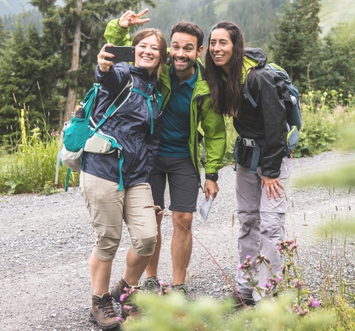 Das Bild zeigt drei fröhliche Wanderer, die auf einem bergigen Wanderweg ein Selfie machen und die gemeinsame Zeit in der Natur geniessen.
Im Bild sind drei Personen zu sehen, zwei Frauen und ein Mann, die sich auf einem Schotterweg in einer bergigen Landschaft befinden. Sie sind offensichtlich beim Wandern, denn sie tragen Rucksäcke und Wanderkleidung. Die Person ganz links, eine Frau mit blonden Haaren, hält ein Smartphone und macht ein Selfie mit ihren beiden Begleitern. Sie trägt eine blaue Regenjacke, beige 3/4-Hosen und braune Wanderschuhe. An ihrem Rucksack ist eine türkise Wasserflasche befestigt. Sie lächelt breit und macht eine V-Geste mit zwei Fingern.
Neben ihr steht ein Mann mit dunklem Haar und Bart, der eine grüne Regenjacke über einem blauen T-Shirt und dunklen Shorts trägt. Er lächelt ebenfalls und schaut in die Kamera. Er hält eine kleine Karte oder ein Notizbuch in der Hand.
Ganz rechts ist eine Frau mit dunklem Haar zu sehen, die eine schwarze Regenjacke über einer grauen Wanderhose trägt. Auch sie lächelt und scheint in das Selfie involviert zu sein. Sie trägt einen schwarzen Rucksack.
Der Hintergrund des Bildes zeigt eine üppig grüne Berglandschaft mit vielen Bäumen und sanften Hügeln. Der Himmel ist bewölkt, was auf ein kühleres Wetter hindeutet, passend zur Kleidung der Wanderer. Im Vordergrund sind vereinzelt blühende lila Disteln zu sehen, die dem Bild eine natürliche und leicht wilde Note verleihen.
Die Atmosphäre des Bildes ist sehr positiv, fröhlich und unbeschwert. Die lächelnden Gesichter und die Selfie-Pose vermitteln ein Gefühl von Freundschaft, gemeinsamer Freude und Abenteuerlust. Die Naturkulisse trägt zu einer friedlichen und erfrischenden Stimmung bei. Es ist offensichtlich, dass die drei Personen ihre gemeinsame Wanderung und die Natur geniessen.