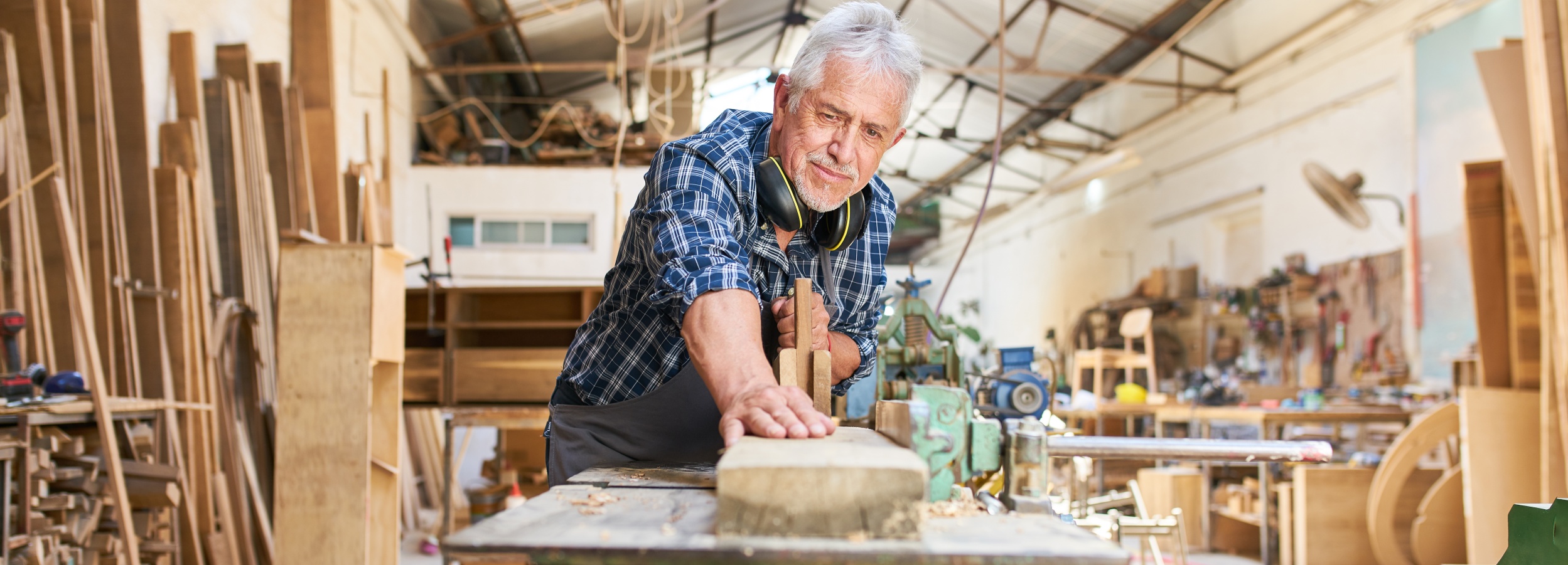 Ein älterer Handwerker arbeitet konzentriert in seiner Werkstatt an einem feinen Holzstück.

Das Bild zeigt einen älteren Mann mit grauem Haar und einem karierten Hemd, der in einer Werkstatt steht und ein Holzstück bearbeitet. Er trägt Kopfhörer um den Hals. Seine Hände sind auf dem Holzstück und einem Stechbeitel platziert. Im Hintergrund sind Regale voller Holzstücke und Werkzeuge zu sehen. Die Werkstatt wirkt belebt und voller Arbeitsmaterialien. Die Atmosphäre ist handwerklich und professionell, geprägt von der Hingabe des Mannes zu seinem Handwerk.