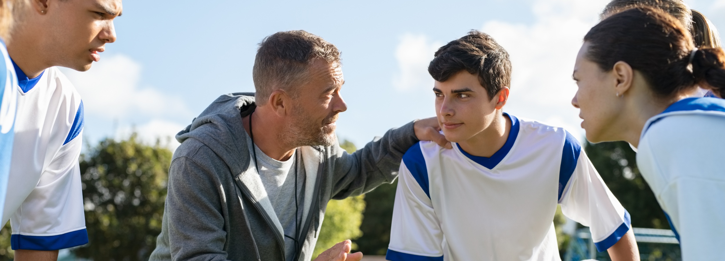 Ein Fußballtrainer gibt seinem Team Anweisungen vor dem Spiel oder in der Halbzeitpause.

Das Bild zeigt einen Fußballtrainer, der mit einigen seiner Spieler spricht. Der Fokus liegt auf dem Trainer, einem Mann mittleren Alters mit grauen Haaren, der eine graue Kapuzenjacke über einem grauen T-Shirt trägt. Er legt seine Hand auf die Schulter eines jungen Spielers, der ihm direkt zuhört. Der Spieler trägt ein weißes Fußballtrikot mit blauen Akzenten an den Schultern und am Kragen.

Um die beiden herum sind weitere Spieler zu sehen, die teilweise angeschnitten sind. Man erkennt auch, dass der Himmel leicht bewölkt ist und Bäume im Hintergrund stehen. Die Atmosphäre wirkt konzentriert und motivierend, da der Trainer seine Weisheit und Strategie mit dem Team teilt. Die Stimmung ist ernst, aber konstruktiv, da alle Beteiligten auf das gemeinsame Ziel fokussiert sind.
