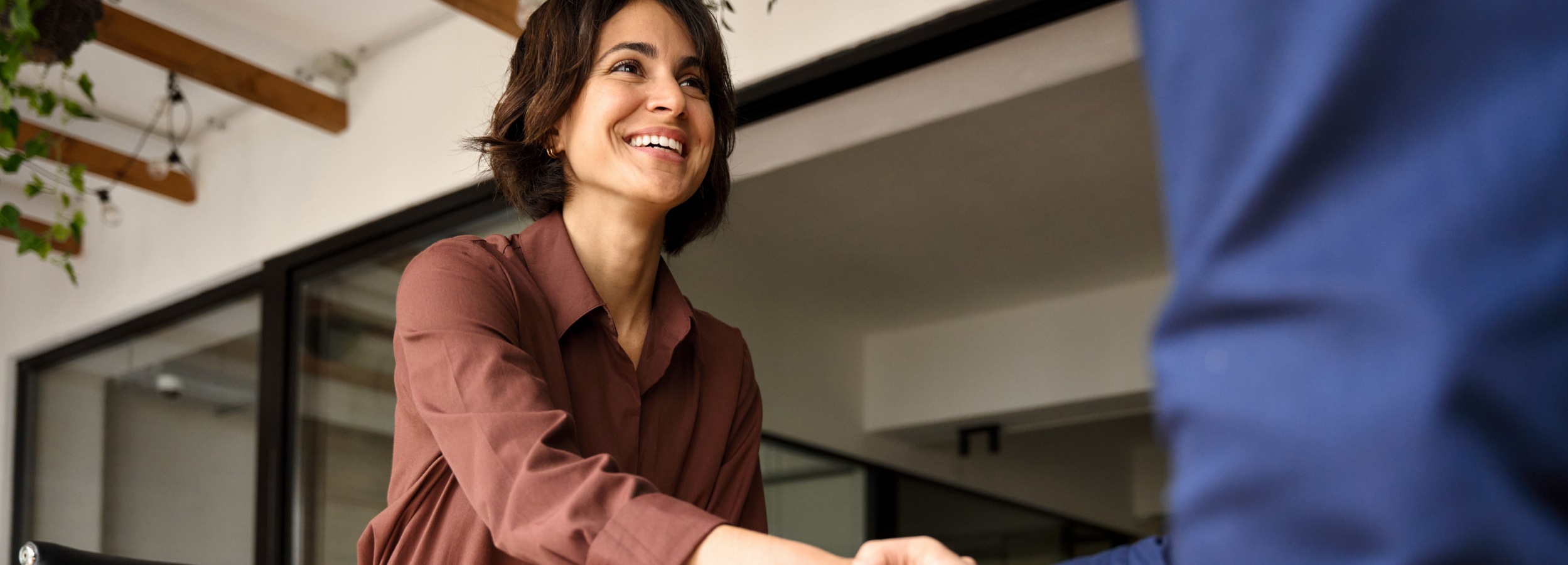 Ein freundliches Lächeln und ein Händedruck signalisieren eine positive geschäftliche Übereinkunft.

Das Bild zeigt eine junge Frau mit dunklem, schulterlangem Haar, die ein burgunderrotes Hemd trägt und herzlich lächelt, während sie scheinbar jemandem die Hand schüttelt. Nur der Arm und ein Teil des Oberkörpers der anderen Person, bekleidet mit einem blauen Jackett, sind im Bild sichtbar. Der Hintergrund ist etwas verschwommen, aber man erkennt Holzbalken an der Decke und eine Fensterfront mit Blick nach draußen, wo grüne Pflanzen hängen. Die Atmosphäre wirkt warm und professionell, und die Stimmung ist positiv und einladend.
