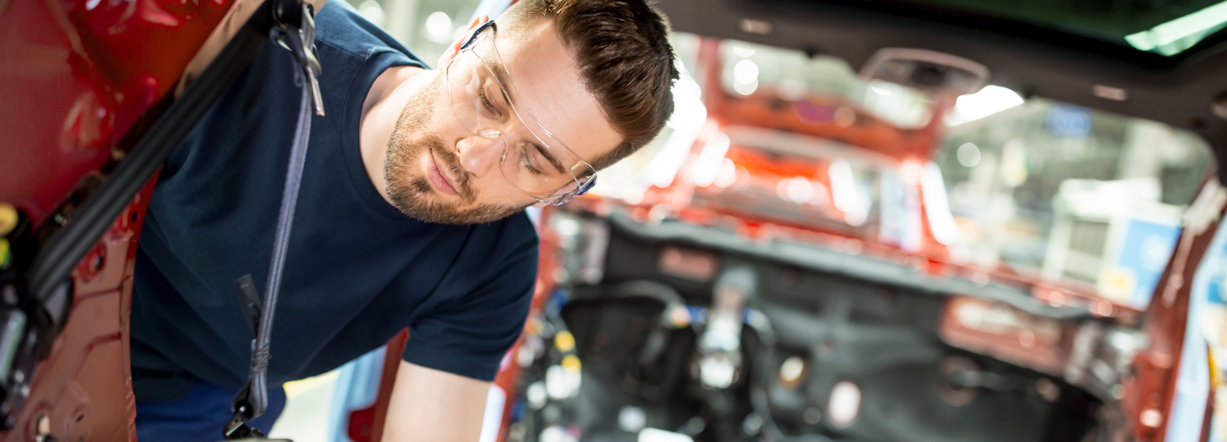 Ein Mann mit Schutzbrille arbeitet konzentriert an der Innenausstattung eines roten Autos. 

Das Bild zeigt einen Mann mit Bart und kurzen braunen Haaren, der eine Schutzbrille trägt und in ein rotes Auto hineinlehnt. Er scheint an der Inneneinrichtung oder Verkabelung des Fahrzeugs zu arbeiten. Sein Gesicht ist nach unten gerichtet, und seine Augen sind auf die Arbeit konzentriert. Er trägt ein dunkelblaues T-Shirt.

Der Hintergrund zeigt das Innere des Autos, das in leuchtendem Rot lackiert ist, sowie verschiedene Kabel und mechanische Teile, die zugänglich sind. Das Sonnenlicht scheint von oben durch ein Schiebedach und beleuchtet den Innenraum. Die Atmosphäre ist hell und professionell, was auf eine Werkstatt oder Produktionsstätte hindeuten könnte.