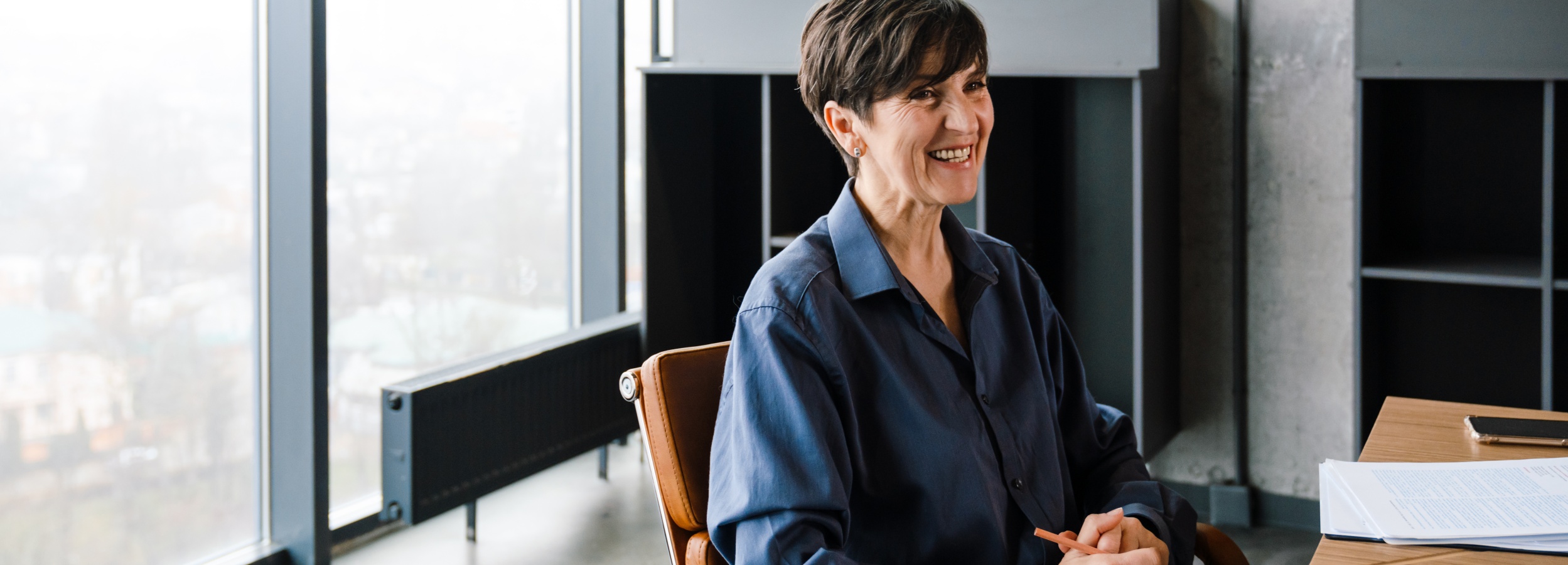 Das Bild zeigt eine lächelnde, reife Frau, die in einem modernen Büro sitzt.

Im Detail zeigt das Bild eine Frau mittleren Alters mit kurzen, dunklen Haaren, die ein dunkles, knielanges Hemd trägt, während sie auf einem braunen Lederstuhl an einem Holztisch sitzt. Ihr Gesicht strahlt ein offenes Lächeln aus, und sie scheint in ein Gespräch vertieft zu sein. Auf dem Tisch vor ihr liegen gestapelte Dokumente, ein Smartphone und ein brauner Bleistift, den sie in ihren Händen hält. Im Hintergrund befindet sich ein großes Fenster, das einen verschwommenen Blick auf eine Stadtlandschaft bietet, sowie ein modernes Regal mit dunklen Fächern. Die Atmosphäre ist hell und professionell, mit einem Hauch von Wärme und Vertrautheit durch das Lächeln der Frau. Die Stimmung wirkt positiv und einladend, was auf ein angenehmes Arbeitsumfeld oder ein entspanntes Gespräch hindeutet.
