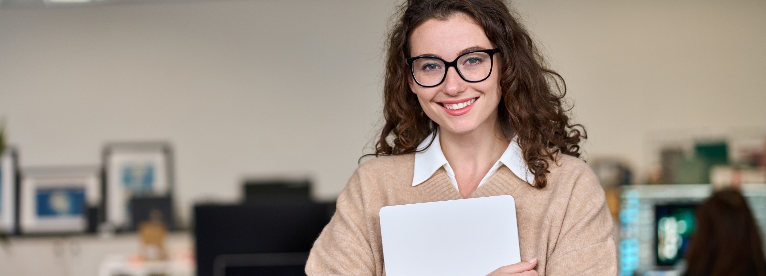 Das Bild zeigt eine lächelnde junge Frau mit Laptop vor einem Bürohintergrund.

Im Detail ist eine junge Frau mit dunklen, lockigen Haaren und Brille zu sehen, die direkt in die Kamera lächelt. Sie trägt eine weiße Bluse unter einem beigen Strickpullover. In ihren Händen hält sie einen weißen Laptop. Im Hintergrund ist ein unscharfer Büroraum erkennbar. Man sieht Schreibtische, Monitore und an der Wand hängende Bilder. Die allgemeine Atmosphäre wirkt professionell und einladend, die Stimmung ist freundlich und positiv.