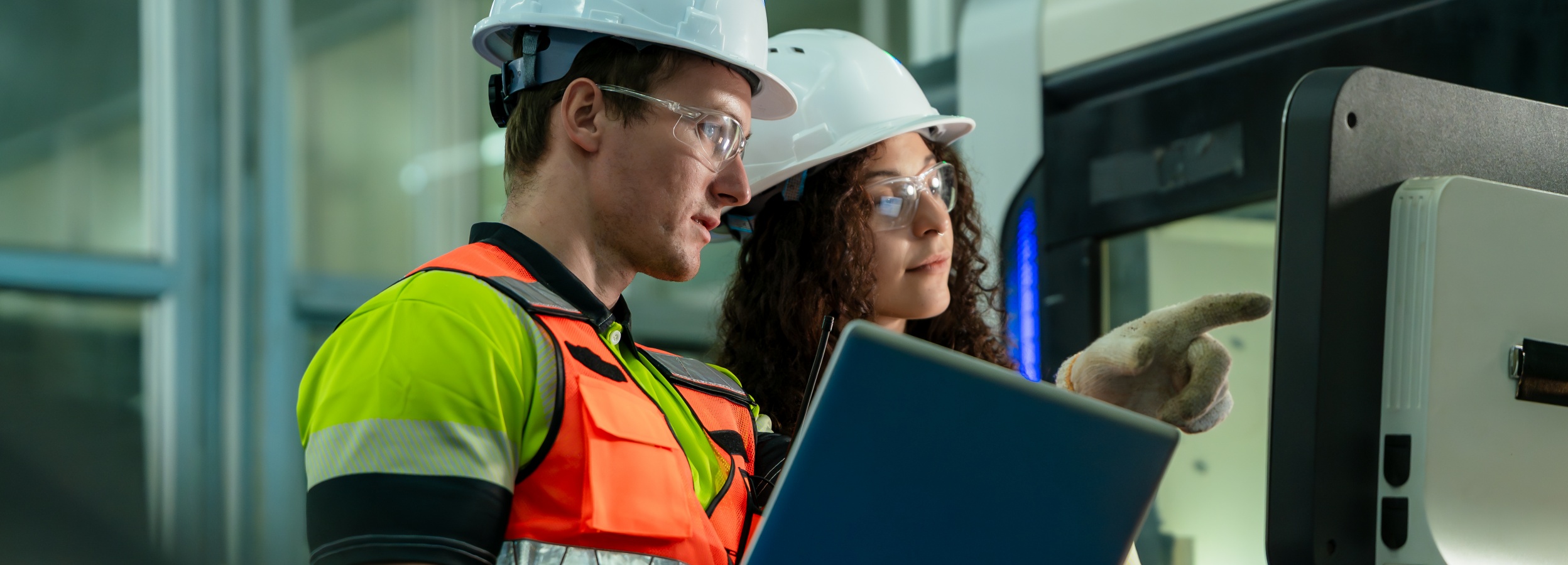 Zwei Ingenieure, ausgestattet mit Sicherheitsausrüstung, arbeiten an einer hochmodernen Maschine, wobei einer auf den Bildschirm deutet und der andere einen Laptop hält.

Das Bild zeigt zwei Personen, vermutlich Ingenieure oder Techniker, die eng zusammenarbeiten. Beide tragen weiße Schutzhelme und Schutzbrillen. Der Mann auf der linken Seite trägt eine auffällige Warnweste in Gelb und Orange über seinem Hemd. Er hat einen Laptop in der Hand. Die Frau auf der rechten Seite hat lockiges Haar und trägt einen Arbeitshandschuh an der Hand, mit der sie auf einen Bildschirm oder eine Maschine deutet.

Hinter den beiden Personen ist ein Teil einer großen, modernen Maschine sichtbar. Der Hintergrund ist unscharf und deutet auf eine industrielle Umgebung hin, möglicherweise eine Fabrik oder Werkshalle. Die Beleuchtung ist hell und gleichmäßig, was eine professionelle und konzentrierte Atmosphäre schafft. Die Stimmung ist arbeitsorientiert und kooperativ, da die beiden Personen in die Arbeit an der Maschine vertieft sind.